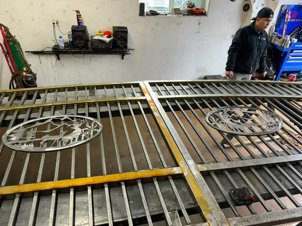 Two steel gate panels with deer scenic medallions on shop table, fabricator working